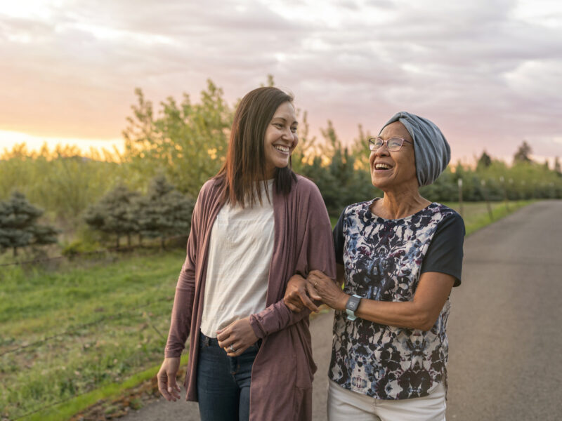 Two women of different ages on a walking path, locked arms and laughing. | Deux femmes d'âges différents sur un sentier de promenade, se tenant par le bras et riant.